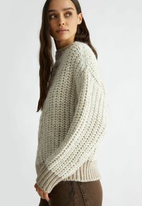 Maglione lavorato a maglia spessa di colore crema con una trama testurizzata e una lavorazione allentata. Presenta un collo alto e polsini a costine. Abbinato a pantaloni marroni.