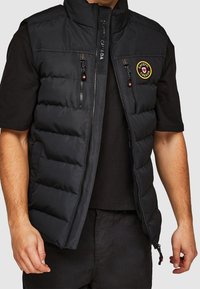 Czarna pikowana kamizelka z wysokim kołnierzem, zamkiem błyskawicznym z przodu i dwoma kieszeniami na zamek. Na lewej piersi znajduje się naszywka z logo.