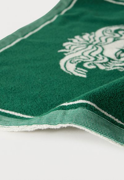 Πράσινη πετσέτα με λευκή μπορντούρα και μερικώς ορατό στυλιζαρισμένο σχέδιο κεφαλής Μέντους πάνω σε λευκή επιφάνεια.