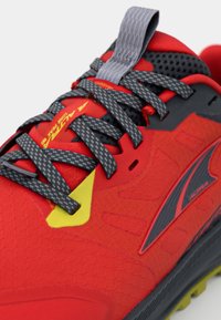 Chaussure de sport rouge avec une tige texturée, des lacets noirs et gris, des accents jaunes et un embout de protection. Caractérisée par une languette rembourrée et un logo.