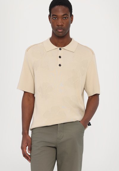 Beige stickad polotröja med ett strukturerat blommönster, tre svarta knappar och korta ärmar, matchad med olivgröna byxor.