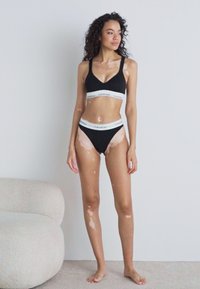 Fekete bralette és bikini szett, fehér Calvin Klein logós derékpánttal. A modell egy semleges színű szőnyegen áll egy sima fal előtt.