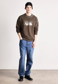 Brun sweatshirt med grafiskt tryck, blå jeans och svarta skor. Modellen bär en svart mössa och visar en avslappnad stil.