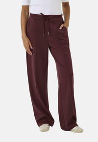 Weinrote Hose mit weitem Bein und Kordelzugbund, zwei Seitentaschen und einem glatten, dehnbaren Stoff für hohen Tragekomfort. Dezente Falten vorne.