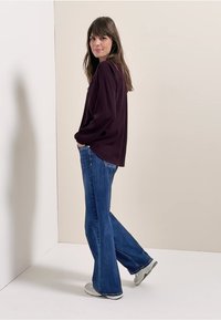 Bordó dlouhý rukáv s volným střihem, doplněný modrými rozšiřujícími se džíny a šedými teniskami, stojící na světlém pozadí.