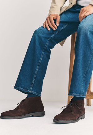Person sitzt auf einem Holzschemel, trägt blaue Jeans und dunkelbraune wildlederne Schnür-Stiefeletten vor hellem Hintergrund.
