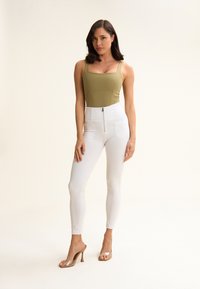Bodysuit verde oliva con escote cuadrado, combinado con pantalones ajustados blancos de cintura alta con cremallera frontal. Zapatos de tacón alto transparentes.