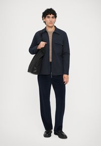 Young man wearing navy jacket, beige sweater, dark pants, black shoes, holding black tote bag on shoulder, standing against plain background.