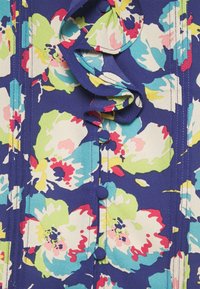 Blomstret stof med livlige blå, grønne, pinke og gule farver, der præsenterer stofbetrukne knapper og en flæse detalje langs midten af sæmsyningen.