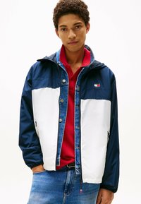 Chaqueta ligera en colores bloqueados azul marino y blanco con detalles en denim, incluyendo capucha, botones a presión y bolsillos laterales.