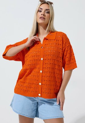 Kvinde med glat blondt hår iført solbriller på hovedet, orange tekstureret skjorte med knapper og lyseblå denimshorts.