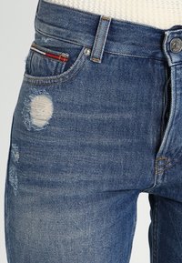 Jeans de mezclilla azul con cintura alta, detalles desgastados y un parche en el muslo izquierdo. Cuenta con un diseño clásico de cinco bolsillos y botón de metal.