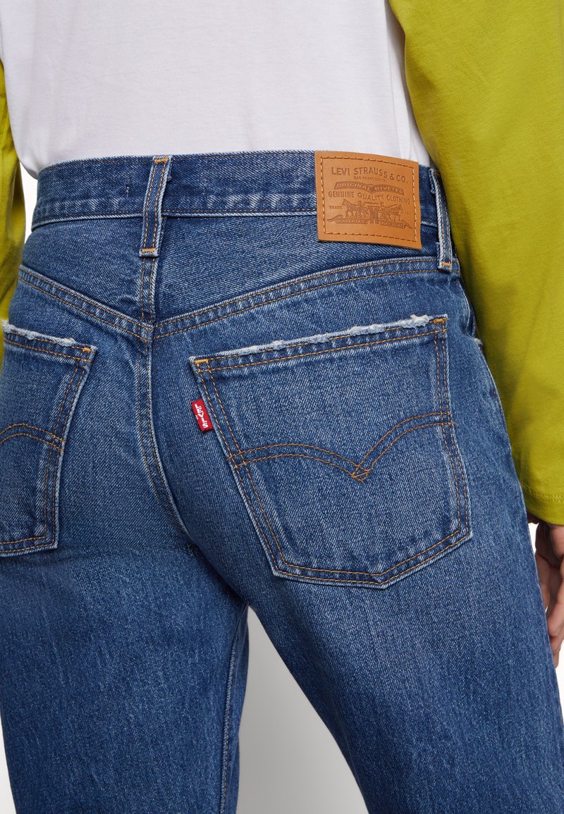 Vaqueros de mezclilla azul con un corte recto, que presentan un parche trasero de cuero, costuras amarillas y una pequeña etiqueta roja de Levi's en el bolsillo.