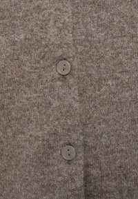 Καφέ πλεκτή ζακέτα με υφή υφάσματος, λεπτομέρεια με κουμπιά και διακριτικό μοτίβο, που παρουσιάζει μια ζεστή, γήινη χρωματική παλέτα.