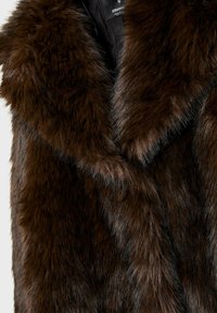 Gilet in pelliccia sintetica marrone con un ampio colletto sovrapposto. La texture è morbida con diverse lunghezze di pelliccia, creando un aspetto stratificato.