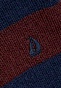 Strickstoff mit diagonalen marineblauen und kastanienbraunen Streifen, der in der Mitte ein kleines marineblaues gesticktes Segelboot-Symbol aufweist.