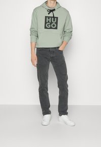 Světle zelená mikina s kapucí a černým logem "HUGO", doplněná tmavě šedými džíny a bílými teniskami. Mikina obsahuje šňůrku na stažení a přední kapsu.