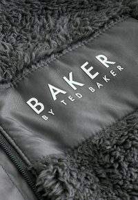 Šedá bunda s fleecovou podšívkou a hladkým vonkajším povrchom. Výrazné logo „BAKER by TED BAKER“ v bielej farbe, umiestnené v strede na tmavom pozadí.