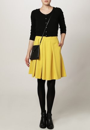 Cardigan noir avec des boutons en perles, jupe plissée jaune, collants noirs et bottines ; comprend un petit sac bandoulière noir.