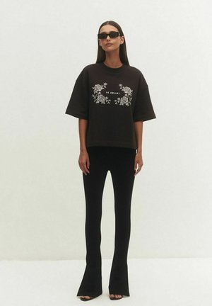 Kobieta nosząca czarne okulary przeciwsłoneczne, czarną oversize koszulkę z białym kwiatowym nadrukiem i napisem „Le Collet”, dopasowane czarne spodnie dzwony oraz buty z odkrytymi palcami.