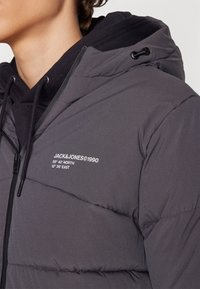 Grå pufferjakke med lynlås foran, med en sort hætte og logo detaljer. Glat tekstur med kviltede sektioner til isolering.