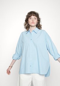 Camisa azul clara com colarinho, mangas enroladas e um corte descontraído. Feita de tecido leve, com uma textura subtil.