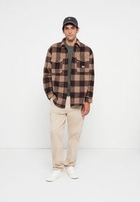 Bruin en tan geblokte flanellen jas, groene shirt, beige broek, witte sneakers en een donkere pet met een klein logo. Eenvoudige, casual outfit.