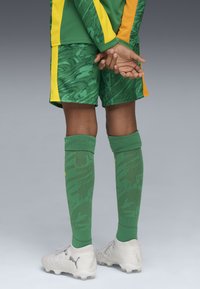 Uniforme deportivo verde y amarillo con pantalones cortos estampados, calcetas verdes hasta la rodilla y zapatillas ligeras grises con superficie texturizada.