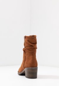 Stivaletti alla caviglia in suede marrone con un design morbido, tacco in legno a strati e dettagli di cucitura sottili lungo il lato. Texture liscia.