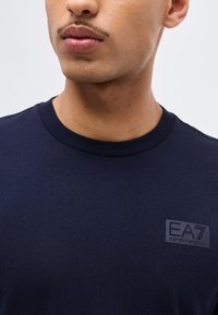 Κοντινό πλάνο ενός ατόμου που φοράει μπλε σκούρο T-shirt EA7 Emporio Armani, με μόλις εμφανή τον λαιμό, τα χείλη και μέρος του προσώπου, σε απλό φόντο.