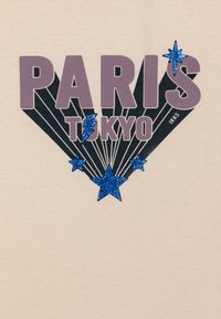 Beige shirt met gedurfde tekst "PARIJS" en "TOKYO", blauwe glittersterren en zwarte grafische lijnen die vanuit het midden stralen.