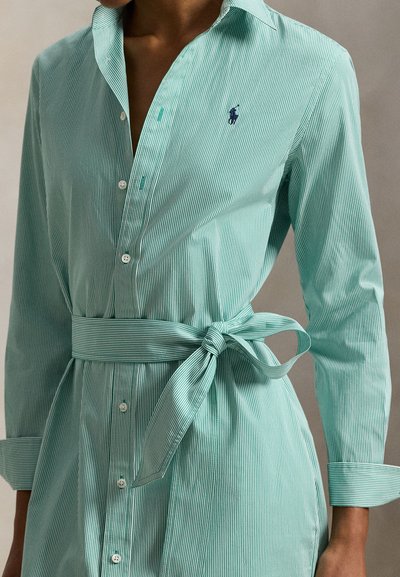 Πουκάμισο με μακρύ μανίκι σε ανοιχτό μπλε και άσπρες ρίγες, με ζώνη στη μέση που δένει και γιακά· διαθέτει κεντημένο λογότυπο στο στήθος.