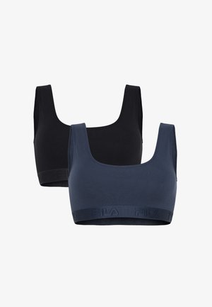 Jonge vrouw staat op blote voeten, draagt een zwarte sportbeha en bijpassende slip tegen een effen lichte achtergrond.