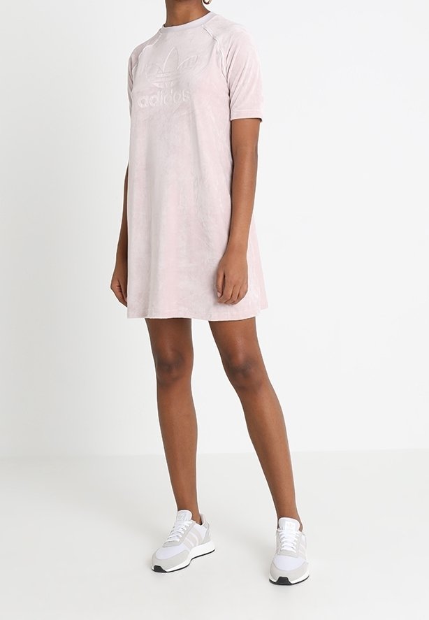Jasnoróżowa sukienka Adidas z krótkimi rękawami, z wybijanym logo i luźnym krojem, zestawiona z białymi i szarymi trampkami. Gładka tekstura.