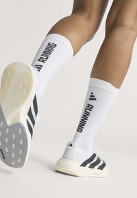 Jambes portant des chaussettes de course blanches avec un texte noir et des baskets blanches à rayures noires marchant sur un fond clair.
