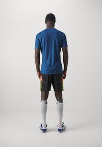 Tricou sportiv albastru, pantaloni scurți negri cu accente neon, șosete albe cu dungi negre și ghete albastre, văzut din spate.