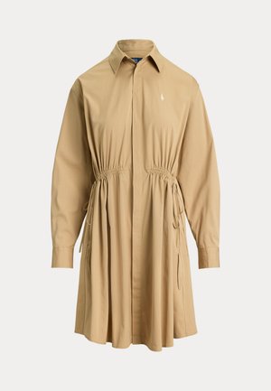 Kobieta w beżowej sukience koszulowej z paskiem w paski, beżowym kapeluszu typu bucket, okularach przeciwsłonecznych, białych trampkach, trzymająca małą pomarańczową torebkę.