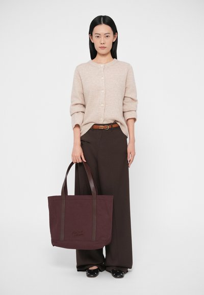 Nagy bordó bordó vászontáska barna bőr fogantyúkkal, tónusú varrással és dombornyomott logóval, bézs kardigánnal és barna nadrággal párosítva.