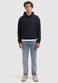 Jonge man met krullend haar, gekleed in een zwarte hoodie met de tekst "PURE PATH", lichte spijkerbroek, beige schoenen, staande tegen een effen achtergrond.