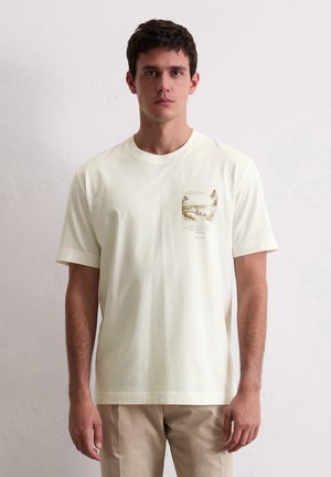 Mand iført en kridhvid T-shirt med et lille brunt naturbillede og tekst på venstre bryst, parret med beige bukser.