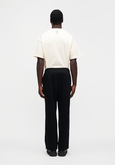 Εκρού κοντομάνικη μπλούζα με πράσινο γραφικό στην πλάτη, συνδυασμένη με μαύρο παντελόνι με ελαστική μέση. Το μοντέλο είναι γυρισμένο πλάτη σε ουδέτερο φόντο.