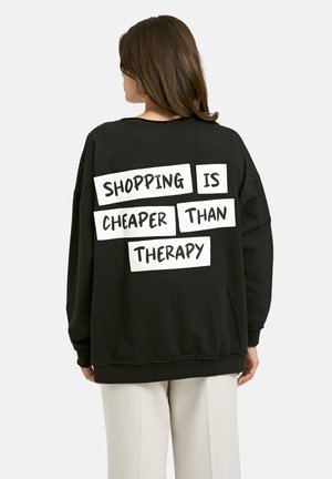 Black sweatshirt with long sleeves. Rear displays large white text: “SHOPPING IS CHEAPER THAN THERAPY” in rectangular frames. Soft texture.