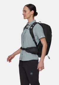 Zwarte rugzak met verstelbare banden en heupriem, gedragen over een lichtgroen geruit shirt. Heeft een logo aan de voorkant en een ergonomisch ontwerp.