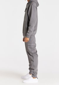 Šedá mikina s kapucí a přední kapsou, se stahovací šňůrkou, spárovaná s odpovídajícími šedými teplákovými kalhotami s bočními kapsami a manžetami. Bílé tenisky.