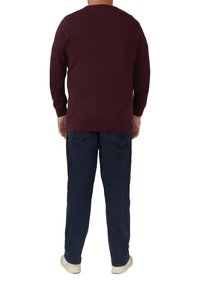 Man die met zijn rug naar de camera staat, gekleed in een maroon sweater, donkerblauwe jeans en witte sneakers tegen een witte achtergrond.