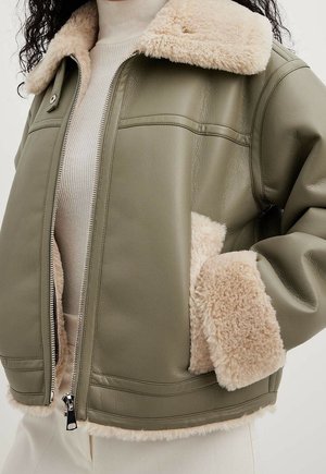 Kurtka ze skóry w oliwkowej zieleni z beżowym kołnierzem z owczej skóry, mankietami i wykończeniem kieszeni, noszona na beżowym golfie.