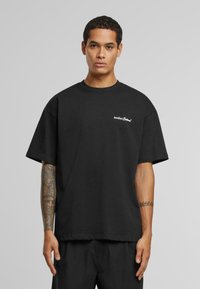 Sort oversized bomulds t-shirt med korte ærmer, med hvid logo tekst på venstre bryst. Glat tekstur, afslappet pasform.