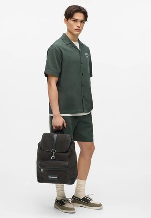 Jauns vīrietis valkā tumši zaļu īsroku kreklu ar pogām un atbilstošas bikses, tur melnu mugursomu, valkā bēša krāsas zeķes un divtoņu laivu apavus.