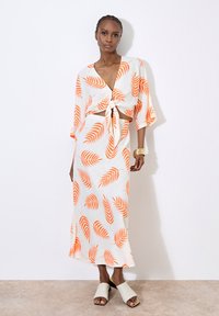 Robe blanche avec des motifs de feuilles oranges, détail à nouer à l'avant, manches larges et silhouette fluide. Associée à des chaussures à enfiler de couleur claire.