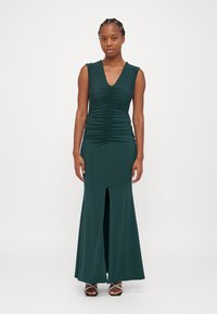 Green sleeveless dress with a V-neck, ruched bodice, and a front slit. Made from smooth fabric, it has a fitted silhouette and flared bottom.